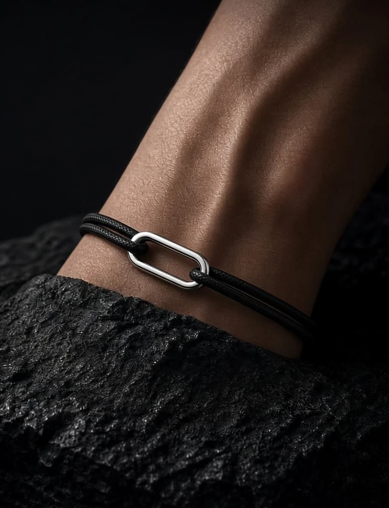 A wrist resting on dark stone, wearing the Pathway: a polished sterling silver link on black braided cord
